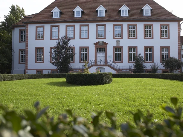 Ein historisches, zweigeschossiges Gebäude mit roten Dachziegeln und weißen Fensterrahmen, umgeben von gepflegten Gärten.