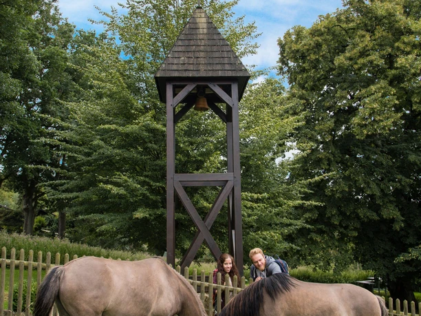 Pferde an der Kirchstelle Andepen Pferde grasen vor einem Holzglockenturm, flankiert von zwei Personen, umgeben von Bäumen.