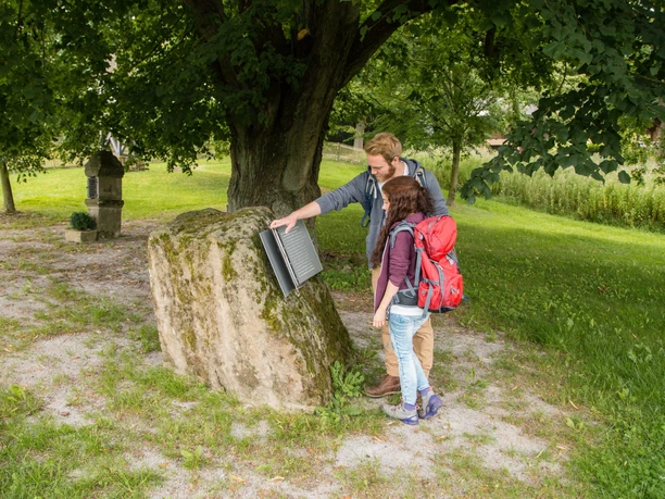 Eisernes Buch Andepen Zwei Menschen betrachten ein offenes, metallisches Buch auf einem Moos-bedeckten Stein unter einem Baum.