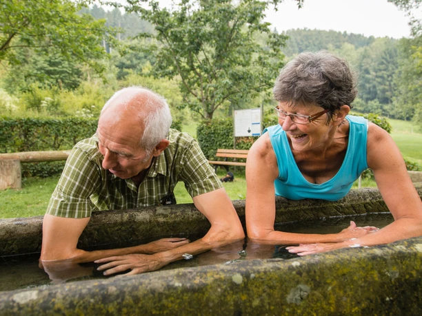 Kneipp-Armbecken im Golmeketal Zwei ältere Menschen erfrischen ihre Arme in einem Kneipp-Armbecken, umgeben von Natur.