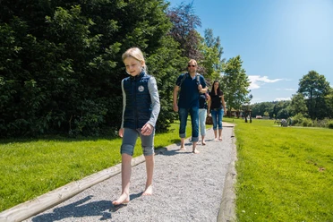 Menschen spazieren barfuß auf einem Kiesweg durch einen grünen Park bei klarem Himmel und Sonnenschein.