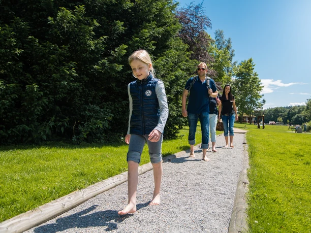 Menschen spazieren barfuß auf einem Kiesweg durch einen grünen Park bei klarem Himmel und Sonnenschein.