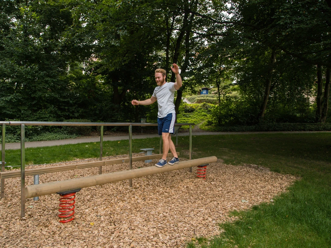 Ein Mann balanciert in einem grünen Park auf einem Holzstamm mit Sprungfedern an den Enden.