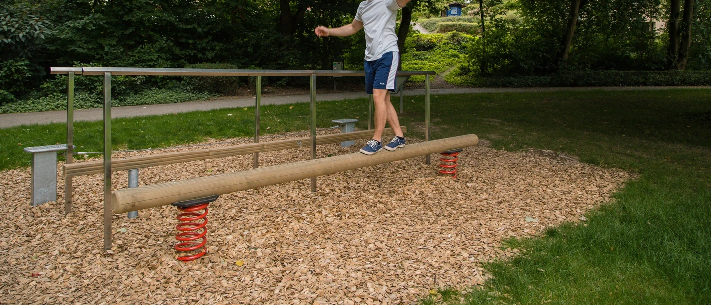 Ein Mann balanciert in einem grünen Park auf einem Holzstamm mit Sprungfedern an den Enden.