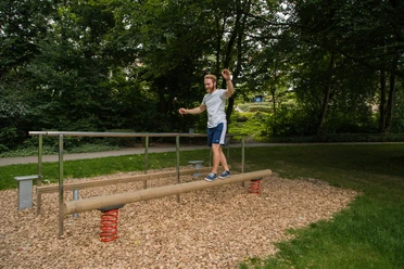 Ein Mann balanciert in einem grünen Park auf einem Holzstamm mit Sprungfedern an den Enden.