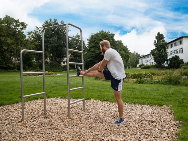 Mann dehnt sein Bein an Sprossenwand auf einer gepflegten Grünfläche unter blauem Himmel im Freien.