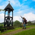 Zwei Wanderer zeigen auf einen hölzernen Glockenturm vor einem Zaun, umgeben von grüner Landschaft.