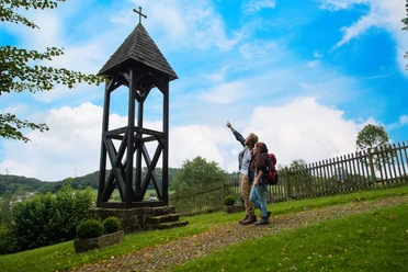 Wanderer bei Andepen Zwei Wanderer zeigen auf einen hölzernen Glockenturm vor einem Zaun, umgeben von grüner Landschaft.