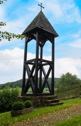 Wanderer bei Andepen Zwei Wanderer zeigen auf einen hölzernen Glockenturm vor einem Zaun, umgeben von grüner Landschaft.
