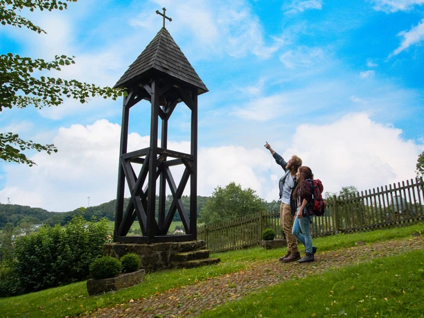 Wanderer bei Andepen Zwei Wanderer zeigen auf einen hölzernen Glockenturm vor einem Zaun, umgeben von grüner Landschaft.
