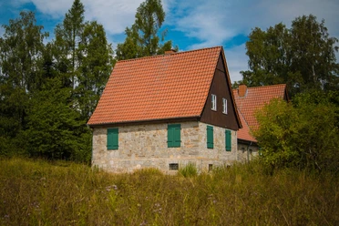 Aahäuser bei Bleiwäsche Ein Steinhaus mit rotem Ziegeldach und grünen Fensterläden, umgeben von Bäumen unter blauem Himmel.