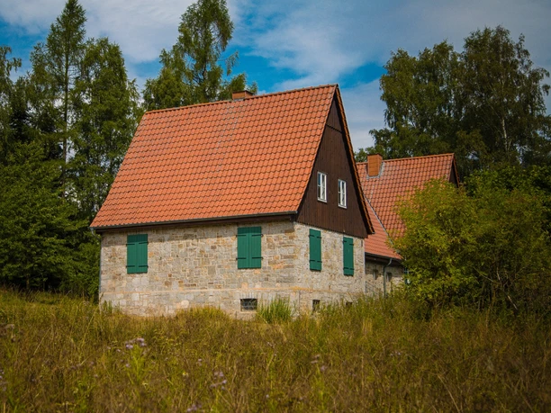 Aahäuser bei Bleiwäsche Ein Steinhaus mit rotem Ziegeldach und grünen Fensterläden, umgeben von Bäumen unter blauem Himmel.