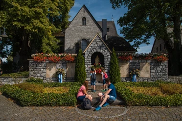 Kirche Bleiwäsche Zwei Erwachsene und drei Kinder sitzen vor einer Steinmauer einer Kirche mit roten Blumen geschmückt.