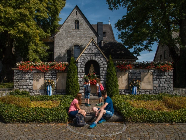 Kirche Bleiwäsche Zwei Erwachsene und drei Kinder sitzen vor einer Steinmauer einer Kirche mit roten Blumen geschmückt.