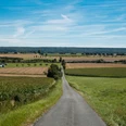 Langer asphaltierter Weg durch weite Felder mit grünem Wiesenrand und blauem Himmel im Hintergrund.