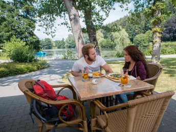 Zwei Personen essen an einem Tisch am See, umgeben von Bäumen und sonniger Landschaft.