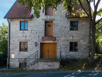 Historische steinerne Mühle mit roten Dachziegeln, umgeben von grünen Bäumen an einem sonnigen Tag.