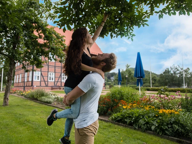 Apfelbaum am Spanckenhof Ein Mann hebt eine Frau, die nach einem Apfel an einem Baum greift, im Garten eines Fachwerkhauses.