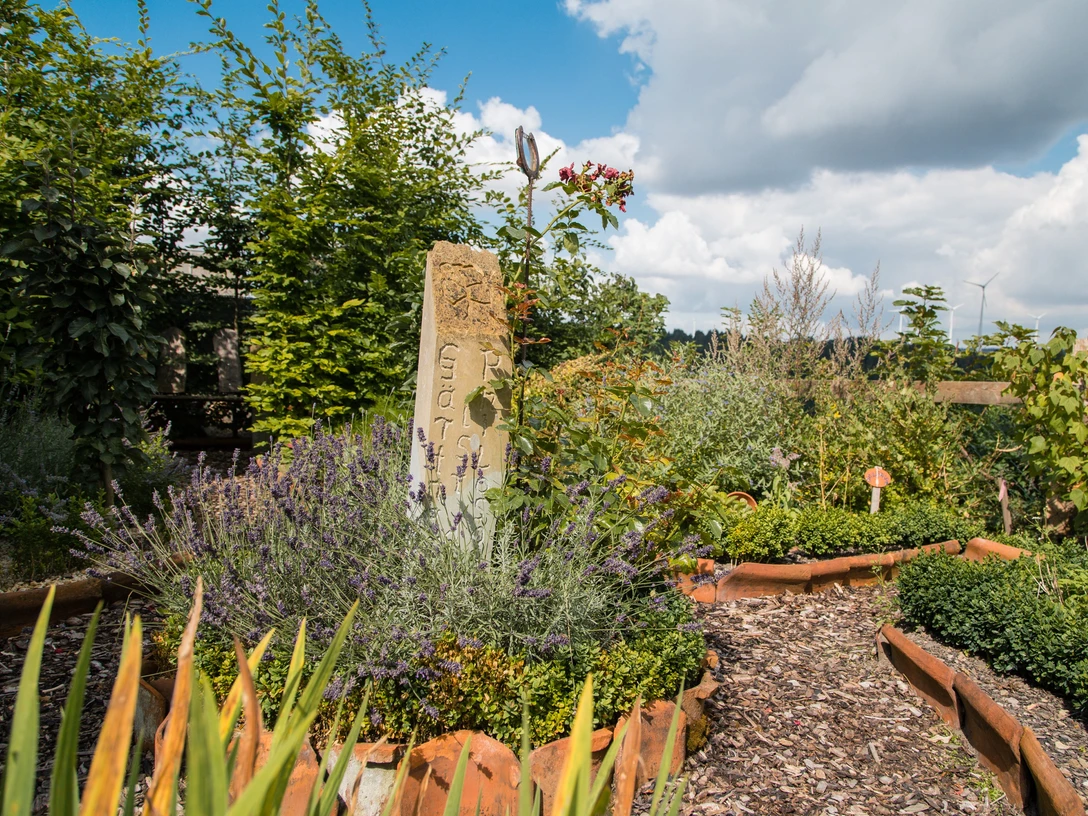 Garten in der Heimatscheune Ein üppiger Garten mit blühenden Pflanzen, gepflasterten Wegen und einem dekorativen Steinsäule.