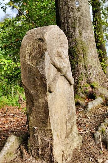 Steinerner Totenschädel-Monolith im Wald, umrahmt von Bäumen und Wurzeln an einem sonnigen Tag.