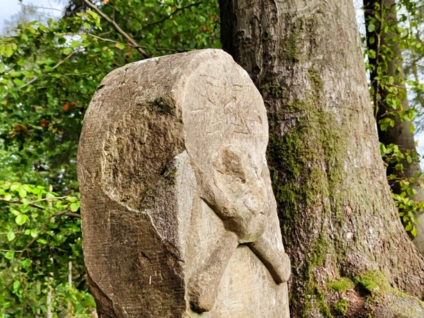 Totenkopfstein Steinerner Totenschädel-Monolith im Wald, umrahmt von Bäumen und Wurzeln an einem sonnigen Tag.