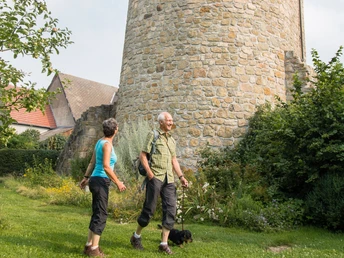 Wandern am Wehrturm Zwei Personen spazieren auf grünem Gras vor einem historischen Wehrturm aus Stein.