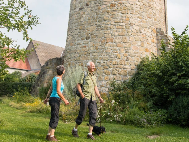 Wandern am Wehrturm Zwei Personen spazieren auf grünem Gras vor einem historischen Wehrturm aus Stein.