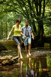 Barfußpfad Bad Wünnenberg Ein Paar spaziert barfuß durch einen flachen Bach, umgeben von üppigem Wald und Bäumen.