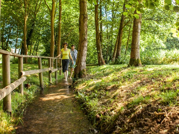 Zwei Personen laufen barfuß auf einem natürlichen Weg durch einen dichten, grünen Waldabschnitt.