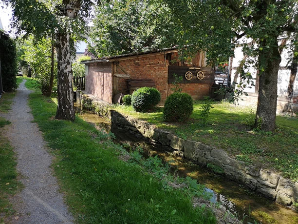 Mühlengraben in Brakel-Bellersen Weg entlang eines kleinen Wassergrabens mit Bäumen und einem Backsteingebäude im Hintergrund.