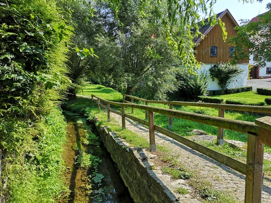 Kleiner Bachlauf mit Holzgeländer, angrenzenden Wiesen und Bäumen, im Hintergrund ein Fachwerkhaus.