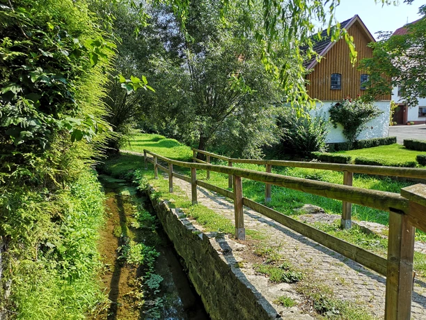Mühlengraben in Brakel-Bellersen Kleiner Bachlauf mit Holzgeländer, angrenzenden Wiesen und Bäumen, im Hintergrund ein Fachwerkhaus.