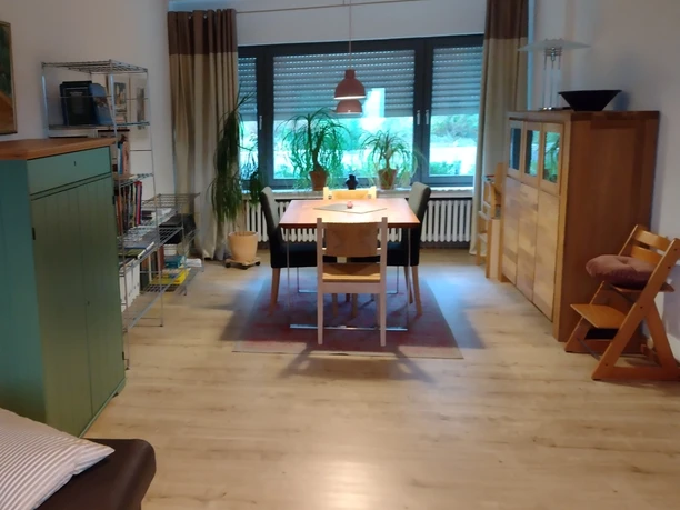 Wohnbereich Ferienwohnung Jakobs Heller Wohnbereich mit Esstisch, Stühlen, Pflanzen und Möbeln aus Holz, große Fenster im Hintergrund.