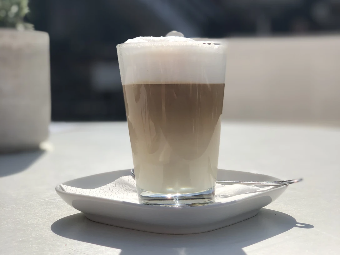 Cappuchino Ein Glas Cappuccino auf einem weißen Tisch im Freien, sonnendurchflutete Szenerie im Hintergrund.