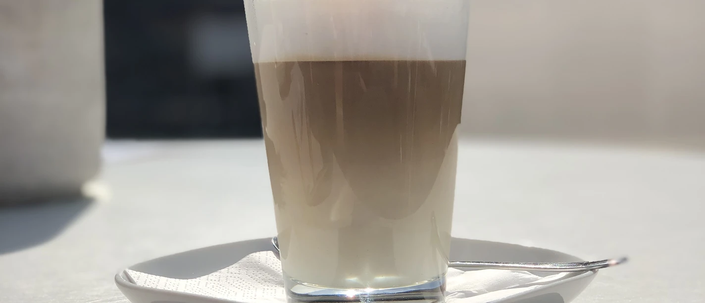 Cappuchino Ein Glas Cappuccino auf einem weißen Tisch im Freien, sonnendurchflutete Szenerie im Hintergrund.