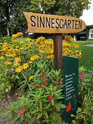 Sinnesgarten in Germete Holzschild für den Sinnesgarten, umgeben von leuchtend gelben und rosafarbenen Blumen.