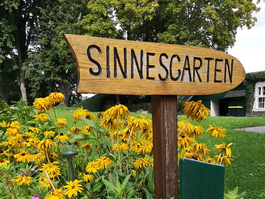Sinnesgarten in Germete Holzschild für den Sinnesgarten, umgeben von leuchtend gelben und rosafarbenen Blumen.
