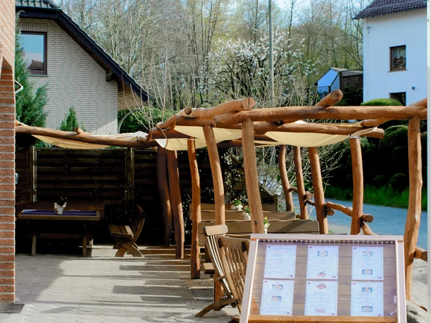 Gasthaus Schadde Biergarten Ein Biergarten mit rustikalen Holzmöbeln und Überdachung, von blühenden Bäumen umgeben, an der Straße.