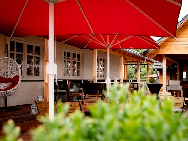 Gemütlicher Biergarten mit roten Sonnenschirmen und hölzernen Sitzmöbeln vor einem Gasthof.