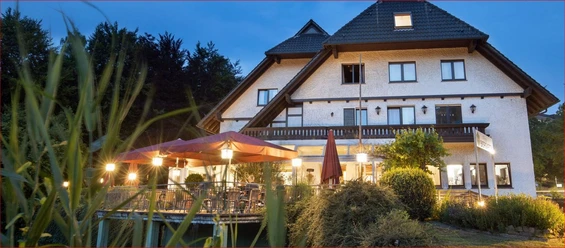 Deekes Quellenhof Ein Landhaus bei Abenddämmerung mit beleuchteter Terrasse und bunten Sonnenschirmen im Vordergrund.