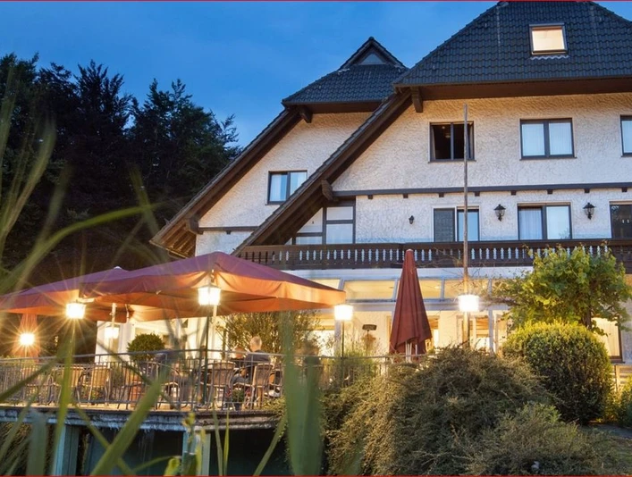 Deekes Quellenhof Ein Landhaus bei Abenddämmerung mit beleuchteter Terrasse und bunten Sonnenschirmen im Vordergrund.