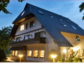 Deekes Quellenhof Großes, gut beleuchtetes Haus mit schrägem Dach, umgeben von üppigem Grün und leuchtenden Lampen.