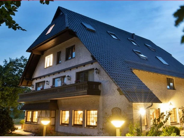 Deekes Quellenhof Großes, gut beleuchtetes Haus mit schrägem Dach, umgeben von üppigem Grün und leuchtenden Lampen.