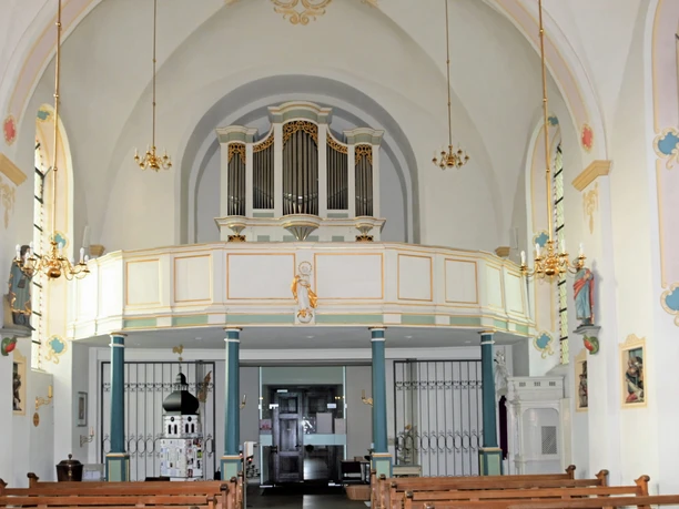 Innenansicht einer Kirche mit bemalten Wänden, Bänken, Orgel auf dem Balkon, und Leuchtern an der Decke.