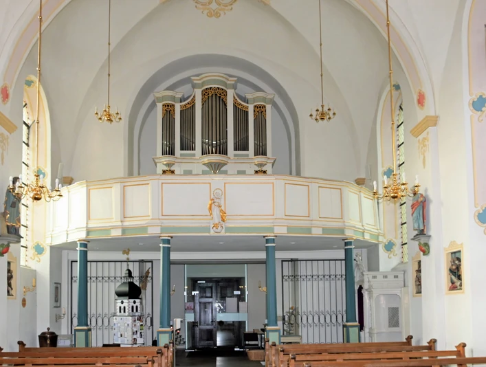 Innenansicht einer Kirche mit bemalten Wänden, Bänken, Orgel auf dem Balkon, und Leuchtern an der Decke.