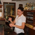 Mittwald Hotel & MCM Restaurant Eine lächelnde Kellnerin zapft ein Getränk an einer Bar, umgeben von Gläsern und Flaschen.