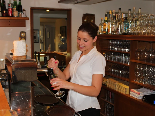 Mittwald Hotel & MCM Restaurant Eine lächelnde Kellnerin zapft ein Getränk an einer Bar, umgeben von Gläsern und Flaschen.