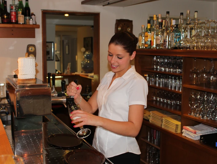 Mittwald Hotel & MCM Restaurant Eine lächelnde Kellnerin zapft ein Getränk an einer Bar, umgeben von Gläsern und Flaschen.