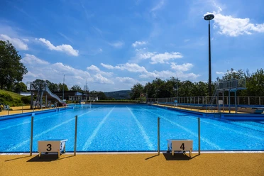 Panoramabad Engelskirchen Freibad mit blauem Schwimmbecken, Sprungbrettern und Rutsche an einem sonnigen Tag mit Wolken am Himmel.