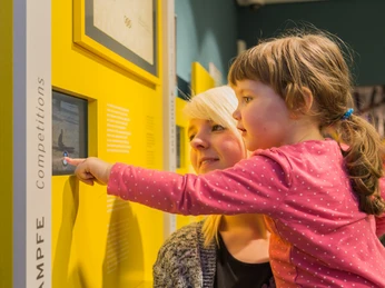Museum Eine Frau mit blondem Haar betrachtet mit einem Kind interaktiv eine Museumsanzeige.A woman with blonde hair interactively looks at a museum display with a child.Žena s blond vlasy si interaktivně prohlíží muzejní expozici s dítětem.Kobieta o blond włosach interaktywnie ogląda wystawę muzealną z dzieckiem.Een vrouw met blond haar kijkt interactief met een kind naar een museumdisplay.Una donna con i capelli biondi osserva in modo interattivo un'esposizione museale con un bambino.